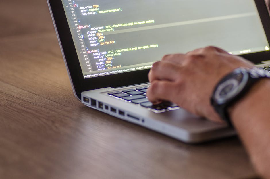 A person is working on a laptop placed on a wooden desk, with their right hand resting on the keyboard. The laptop screen displays lines of code with colorful syntax highlighting, indicating programmi